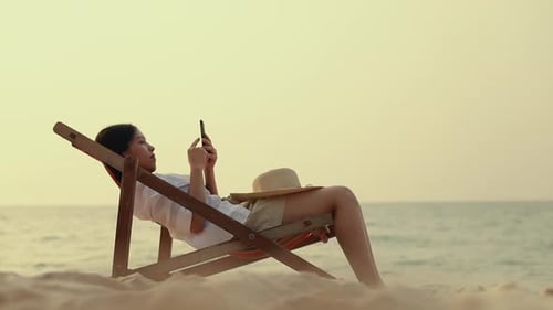 Young Asian woman feeling relax lying on the deck chair on the beach and using smartphone.