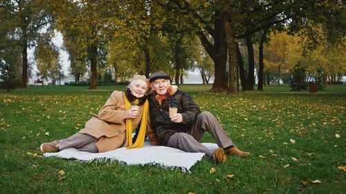 Elderly Husband and Wife Holding Paper Cups of Coffee Smiling Sitting on a Coverlet on Green Grass