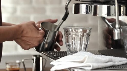 Barista working on coffee maker machine steaming milk in the pitcher in cafe