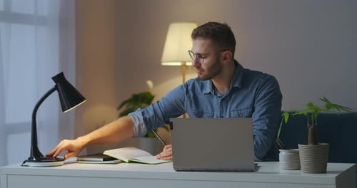 Handsome Male Student Is Learning Online at Evening Writing in Notebook and Searching Info in Laptop