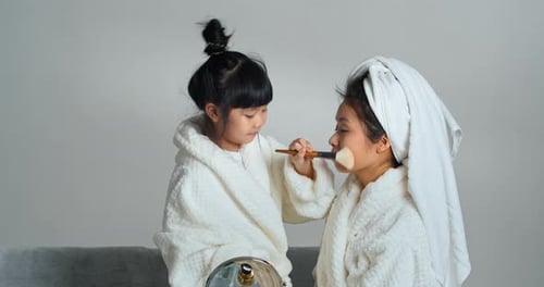 Jolie petite fille ethnique en peignoir blanc assise à la maison avec une maman aimante avec une serviette sur la tête joue
