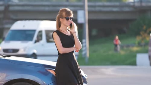 Young Female Driver Standing Near Her Car Talking on Mobile Phone on a City Street in Summer