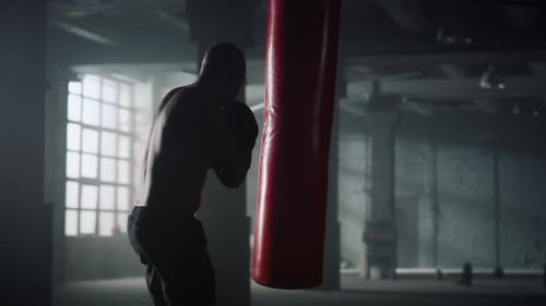 Muscular Man Boxing with Heavy Bag in Dark Gym