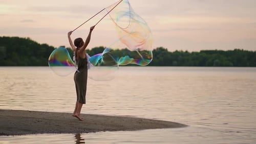 A Young Female Artist Shows a Soap Bubble Show Blowing Up Huge Soap Bubbles on the Shore of a Lake