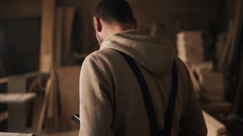 Backside View of a Young Man in a Sweatshirt and Blue Overalls Going Through a Carpentry Workshop