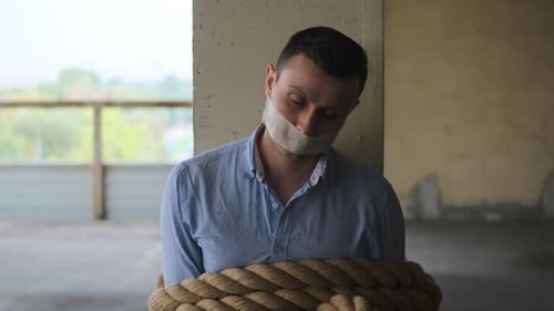 Businessman Tied with Rope with His Mouth Sealed with Tape Sits Tied to Post Looks Up at Camera