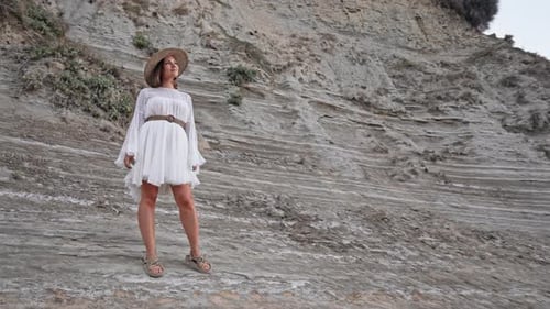 Portrait of Young Woman in White Dress Rope Trendy Sandals and Straw Hat Posing on Clay Beach