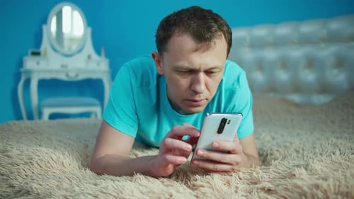 Man Lies in the Bedroom on the Bed with a Mobile Phone in His Hands
