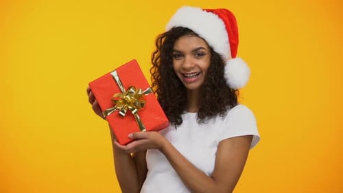 Cheerful Mixed-Race Woman in Santa Claus Hat Holding Red Giftbox and Smiling