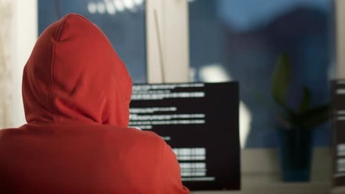 Ein Mann mit Kapuze arbeitet abends zu Hause hinter einem Computerbildschirm, Rückansicht