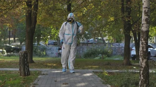 Virologist in a White Protective Suit Disinfects Paving Slabs in a Public Park. The Man Uses a High