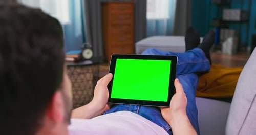 Young Man at Home Laying on a Couch Using with Green Mockup Screen Tablet Computer