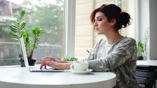 Young Beautiful Woman Sitting Near the Window in Cafe and Working on Laptop Computer