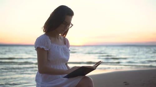 A Young Woman Sits on a Stone on the Beach By the Sea with a Book in Her Hands. A Girl in a White