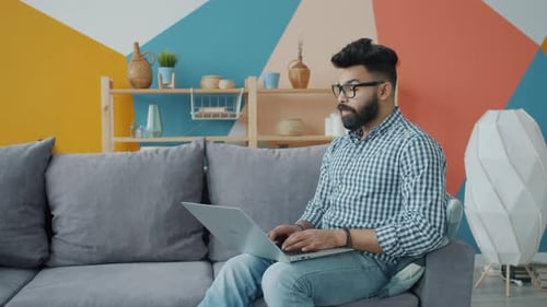 Mixed Race Guy in Glasses and Casual Clothing Using Laptop on Sofa in Apartment