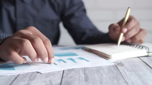 Mão de homem com caneta analisando gráfico de barras em papel e escrevendo no bloco de notas