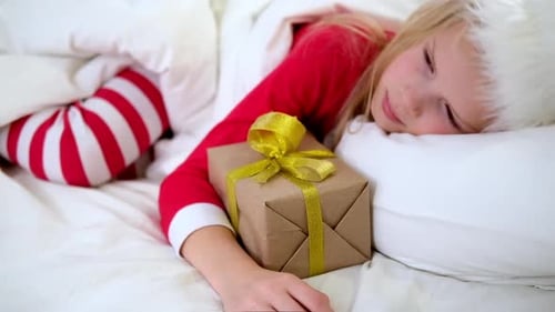 Menina de chapéu de Papai Noel e pijama de Natal deitada na cama com caixa de presente em casa
