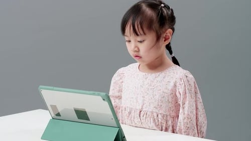 Child using digital tablet sitting near the table to watch cartoon. Record held in studio with gray