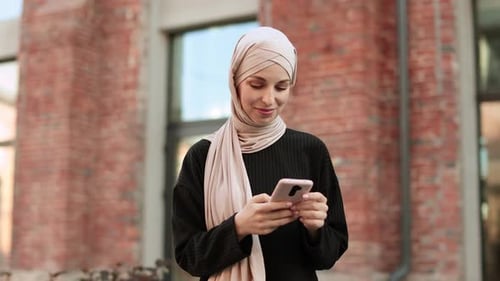 Smiling Muslim Woman Wearing Trendy Black Suit Walking in City Street and Uses Her Phone