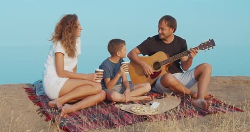Family Sitting By the on the Beach in the Evening and Playing Guitar
