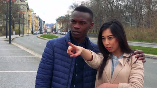 A Young Black Man and a Young Asian Woman Look and Point at Something Off the Camera on a Sidewalk