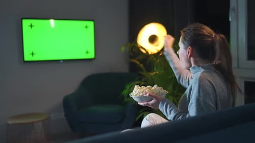 Woman Sitting on a Sofa in the Living Room in the Evening and Watching a Green TV Screen Mockup is