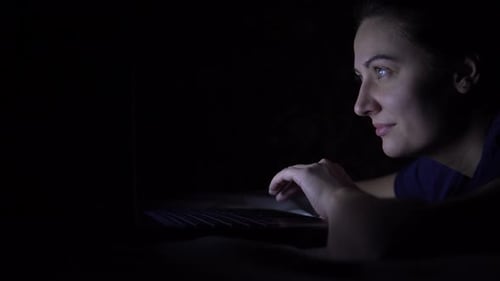 Attractive Happy Woman Typing on a Laptop Keyboard While Lying on a Bed at Night. Isolated Light