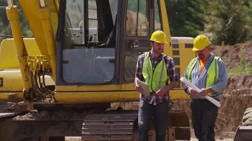 Two workers by excavation equipment look at blueprints