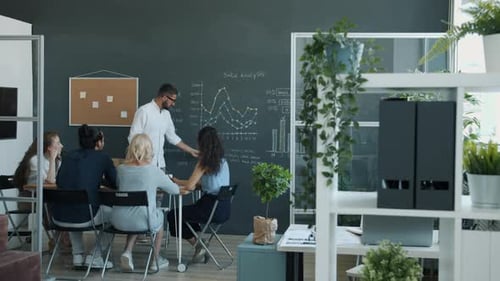 Young Businessman Making Presentation with Chalkboard Speaking to Group of Coworkers in Office