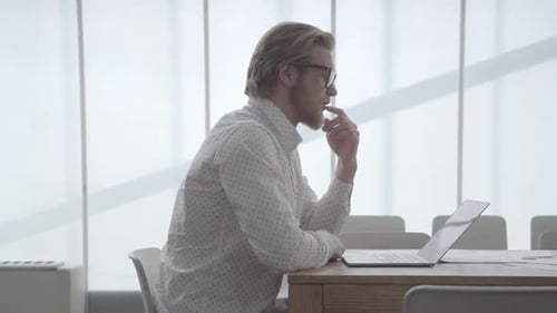 Blond Thoughtful Man in Glasses Sitting at the Table in a Light Comfortable Office