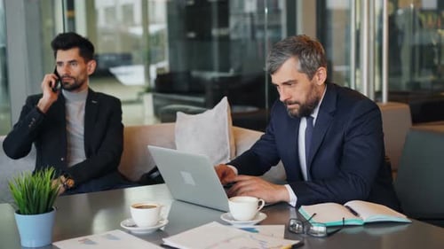 Businessman Using Laptop in Cafe While Partner Talking on Mobile Phone
