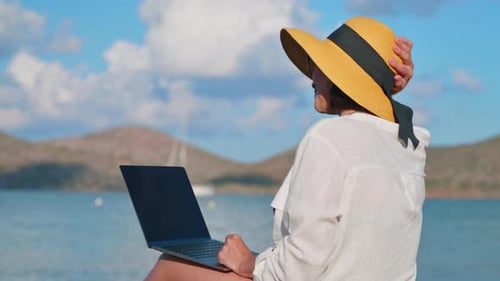 Reife Frau mit Hut mit Laptop am Meeresstrand