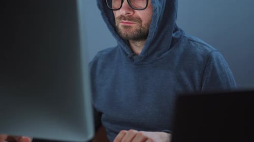 Male Hacker in the Hood and Glasses Working on a Computer in a Dark Office Room. Cybercrime Concept