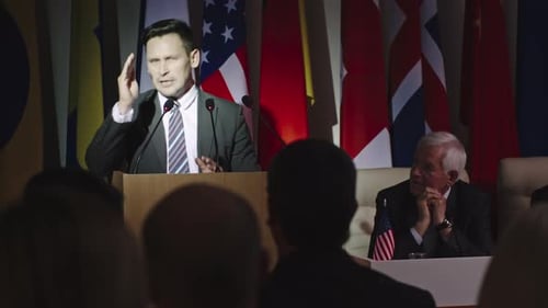 Man Speaks at Podium with Flags Behind Him