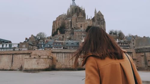 Cinematic Rear View on Stylish Blogger Woman Walking To Stunning Mont Saint Michel Castle Landmark