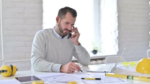 Bearded Man at Desk Talking on Phone About Blueprints