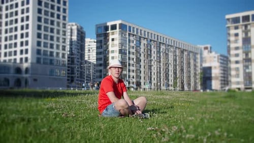 Man in a Hat Sitting on the Grass in the Park on a Background of the Urban Camera Movement