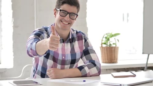 Thumbs Up By Young Man in Office Looking at Camera