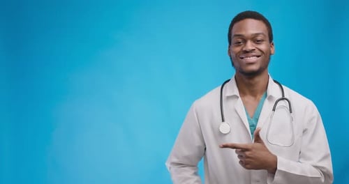 Cheerful African American Doctor Pointing Aside at Copy Space and Smiling, Blue Studio Background