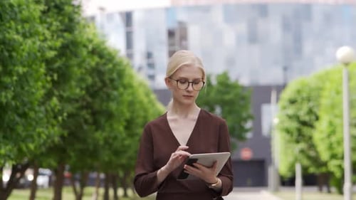 Busy Serious Young Woman with Glasses Walks Down the Street and Scrolls the Tablet