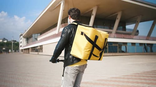 Vista posterior del repartidor con mochila amarilla y máscara protectora montando scooter eléctrico con comida