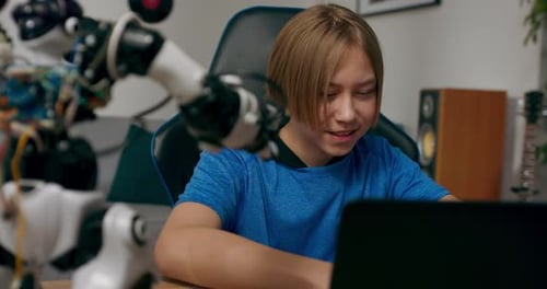 Smart Smiling Ambitious Boy Sits at Desk in Front of Laptop Looking at Robot He is Testing Teenager