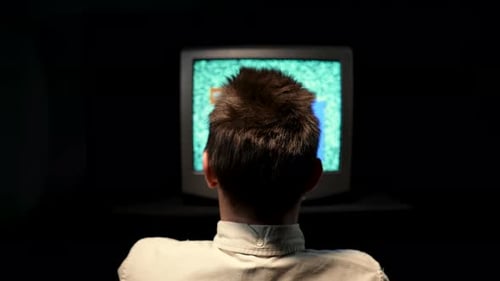 Rear View of a Man Sitting in Front of an Old TV in a Dark Studio on a Black Background