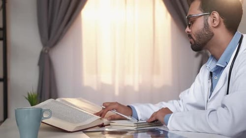 Doctor Writing Notes Next to Open Medical Textbook