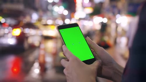 Female tourist holding smart phone with green screen display while standing on the street at night.