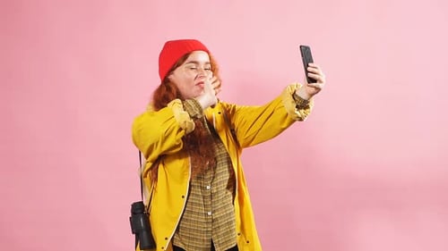 Studio Portrait of a Tourist Girl in a Yellow Raincoat Blowing a Kiss To a Friend on a Smartphone.