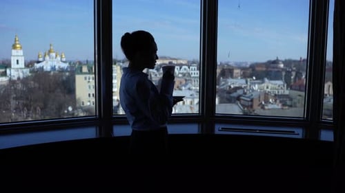 Woman Drinking Coffee Overlooking City From High-Rise Office