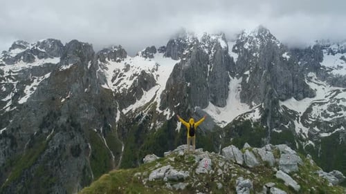 A Hiker Stands on Top of a Mountain and Looks at Snow-capped Mountains, Raising His Hands in Victory