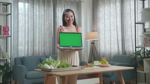 Smiling Asian Woman Showing Green Screen Laptop To Camera While Preparing Healthy Food At Home