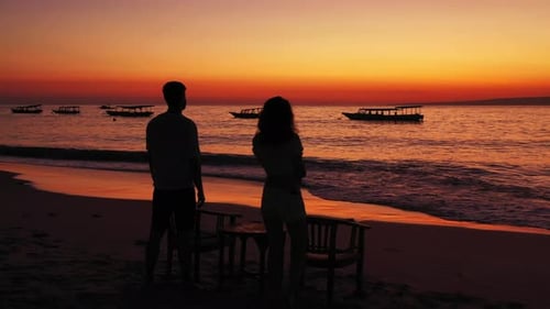 Silhouette of young couple watching romantic sunset with orange sky over sea surface with boats floa